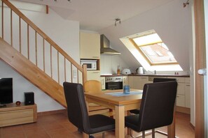 A dining area with a wooden table and chairs, a TV on a stand, and a kitchen in the background. - Holiday apartment with 2 bedrooms (Großenbrode)