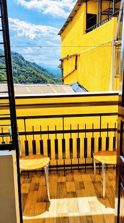 A balcony with a yellow wall, two wooden chairs, and a view of a mountainous landscape.