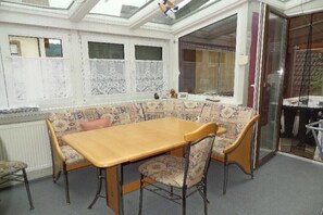 Ein Zimmer mit Holztisch und gemusterten Stühlen, einem Erkerfenster mit Spitzenvorhängen und Blick durch eine Glastür.