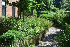 Ein Gartenweg mit Holzzaun, dichter Bepflanzung und einem Gebäude mit Fenstern im Hintergrund.