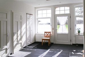 Ein Flur mit weißen Türen, einem hölzernen Stuhl und einem großen Fenster mit Blick auf eine grüne Landschaft.