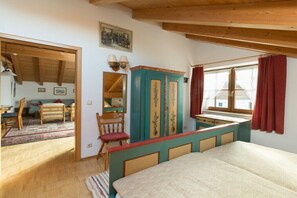 Ein Schlafzimmer mit Bett, Schreibtisch, Stuhl, einem Fenster mit Vorhängen und einer Holzbalkendecke.