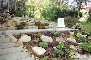 Eine Steintreppe führt zu einem weißen Stuhl, umgeben von einem Garten mit verschiedenen Pflanzen und einer Steinmauer.