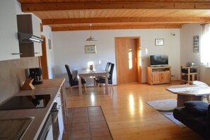Ein Wohnzimmer mit Fernseher, Holzmöbeln, einer Essecke mit Stühlen und einer Küche mit Geräte.