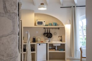 Eine kleine Küche mit einer Mikrowelle, einem Wasserkocher und einem Spülbecken, einem kleinen Kühlschrank sowie einem Fenster mit Vorhängen.