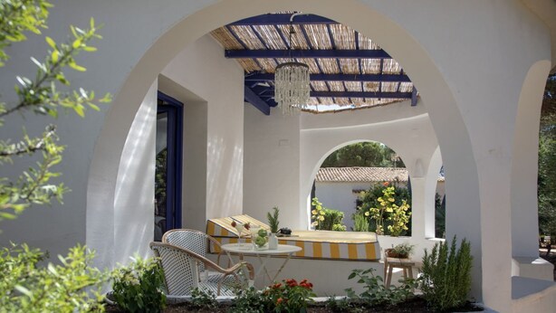 Uma área de estar externa com mesa e cadeiras, cercada por plantas e um guarda-sol decorativo.