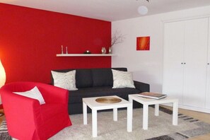 Ein Wohnzimmer mit roter Wand, einer schwarzen Couch, weißen Kissen, einem roten Sessel und zwei weißen Tischen mit Büchern.