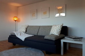 A modern living room with a dark sofa, patterned pillows, a white throw blanket, a floor lamp, a white side table with a book, and three framed artworks on the wall. - Holiday apartment with 1 bedroom (Königstein im Taunus)