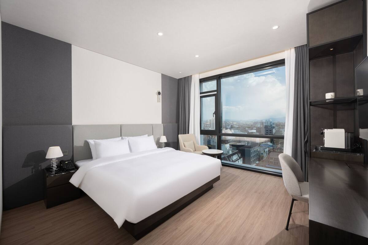 Una habitación de hotel moderna con una cama grande, un escritorio con lámpara, una silla y vistas a la ciudad a través de un ventanal amplio.