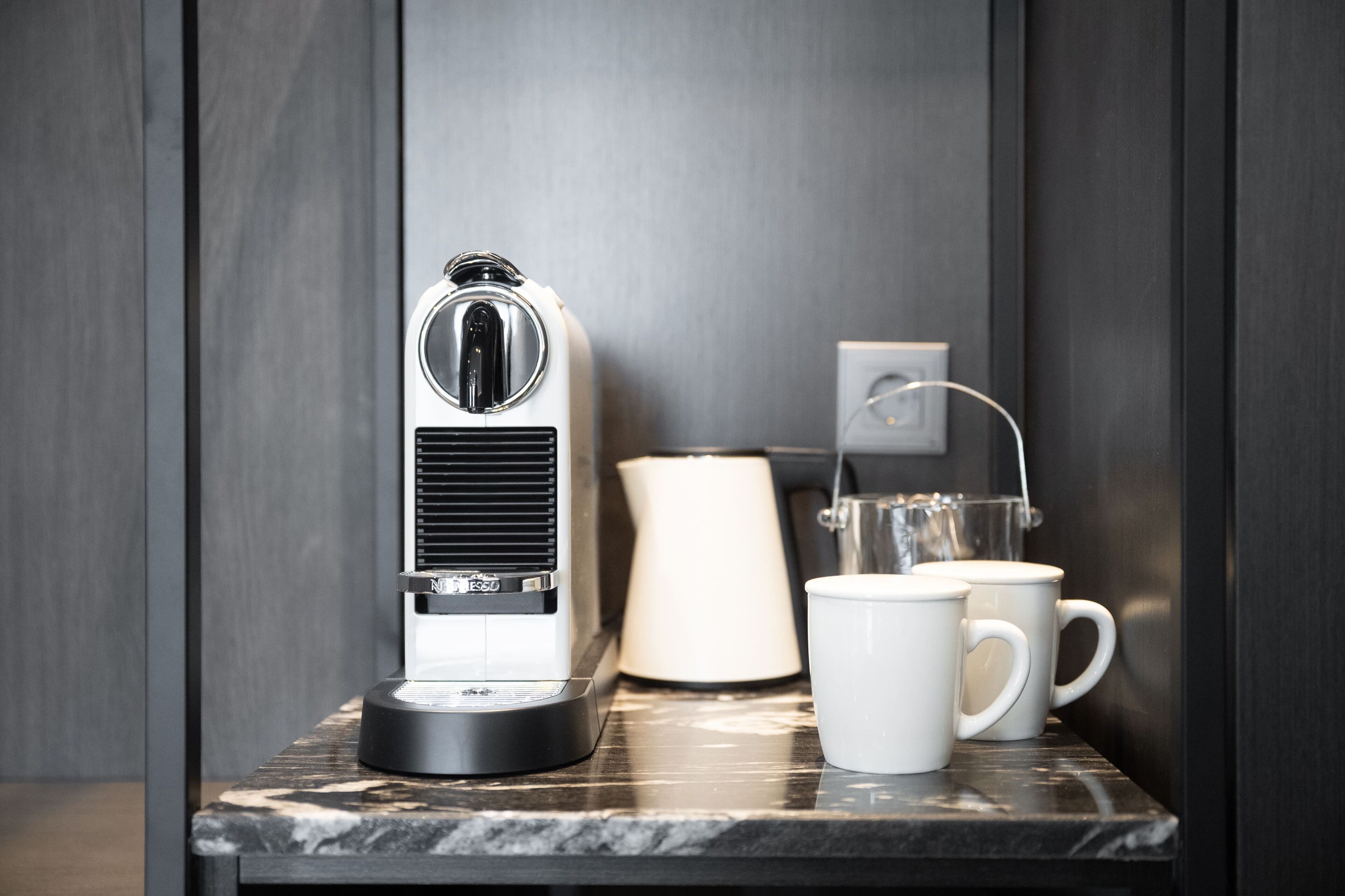 A coffee maker, a lamp, and two white mugs on a marble countertop.