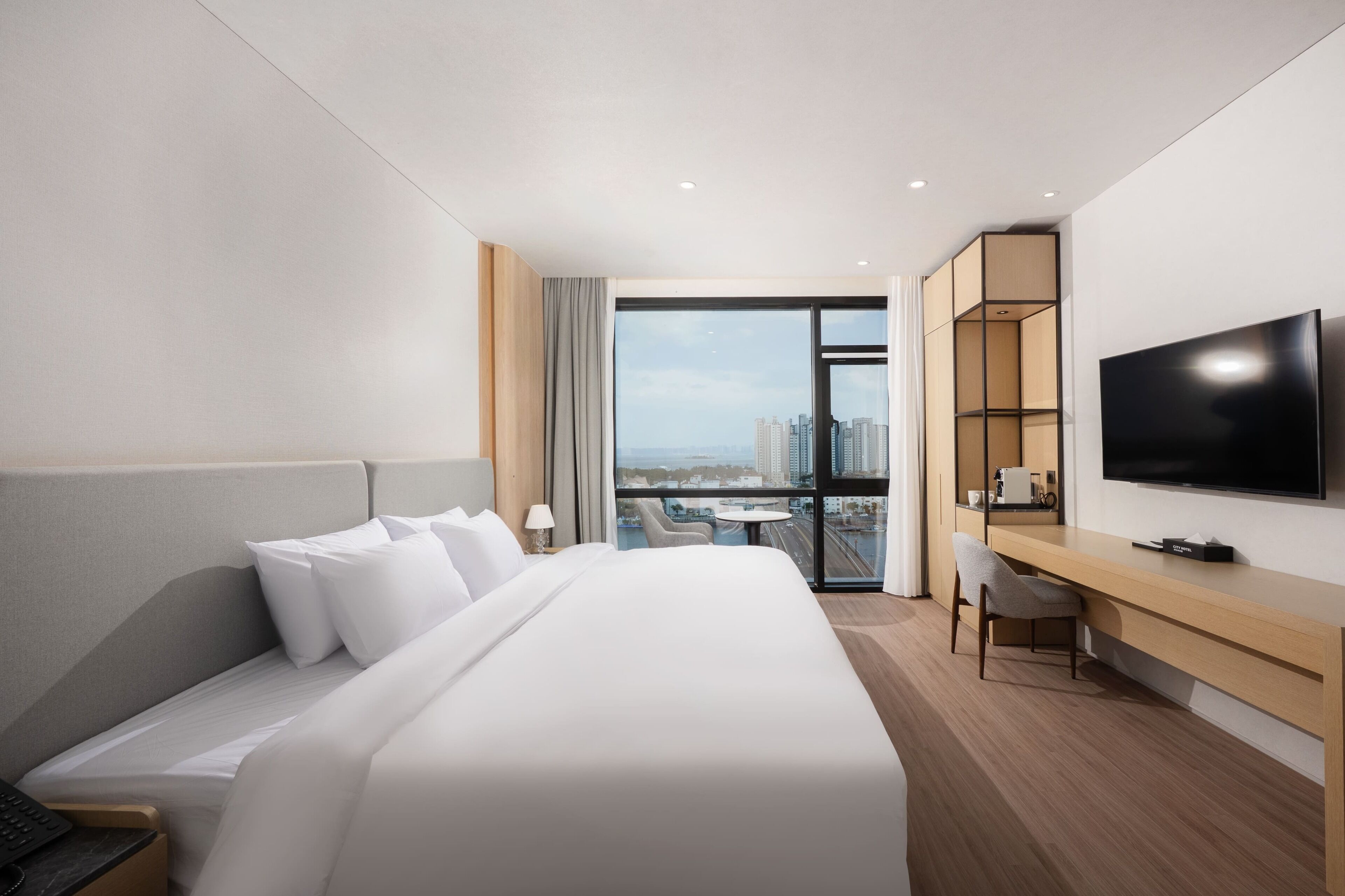 A hotel room with a bed, desk, TV, and a view of the city.