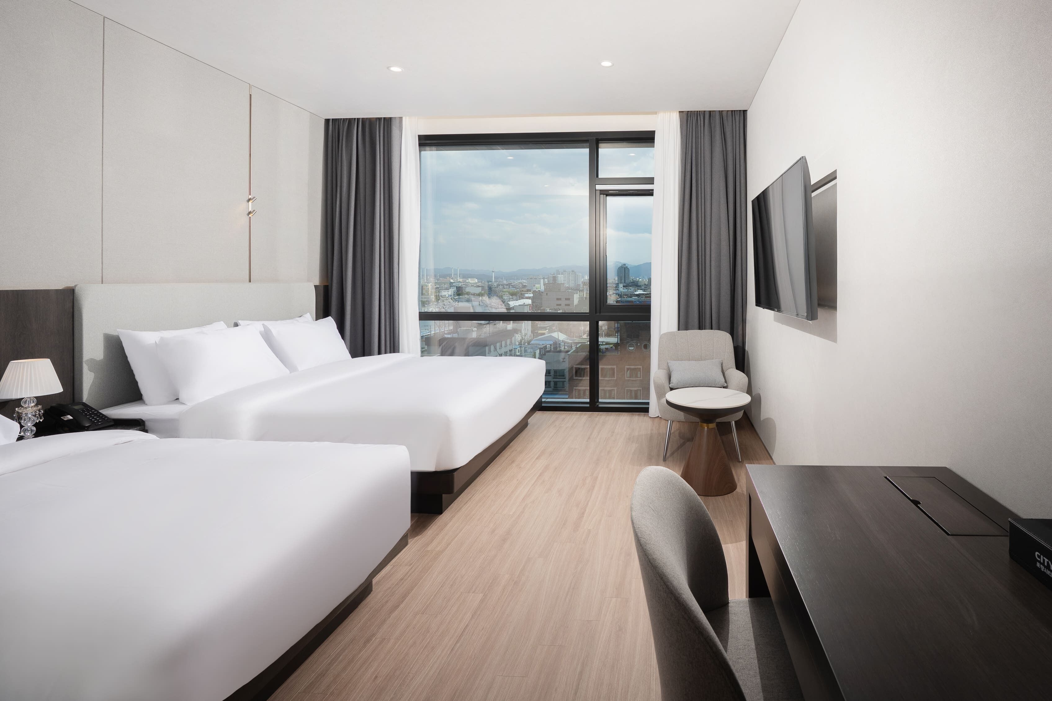 A modern hotel room with two beds, a desk, a chair, a TV, and a large window offering a city view.