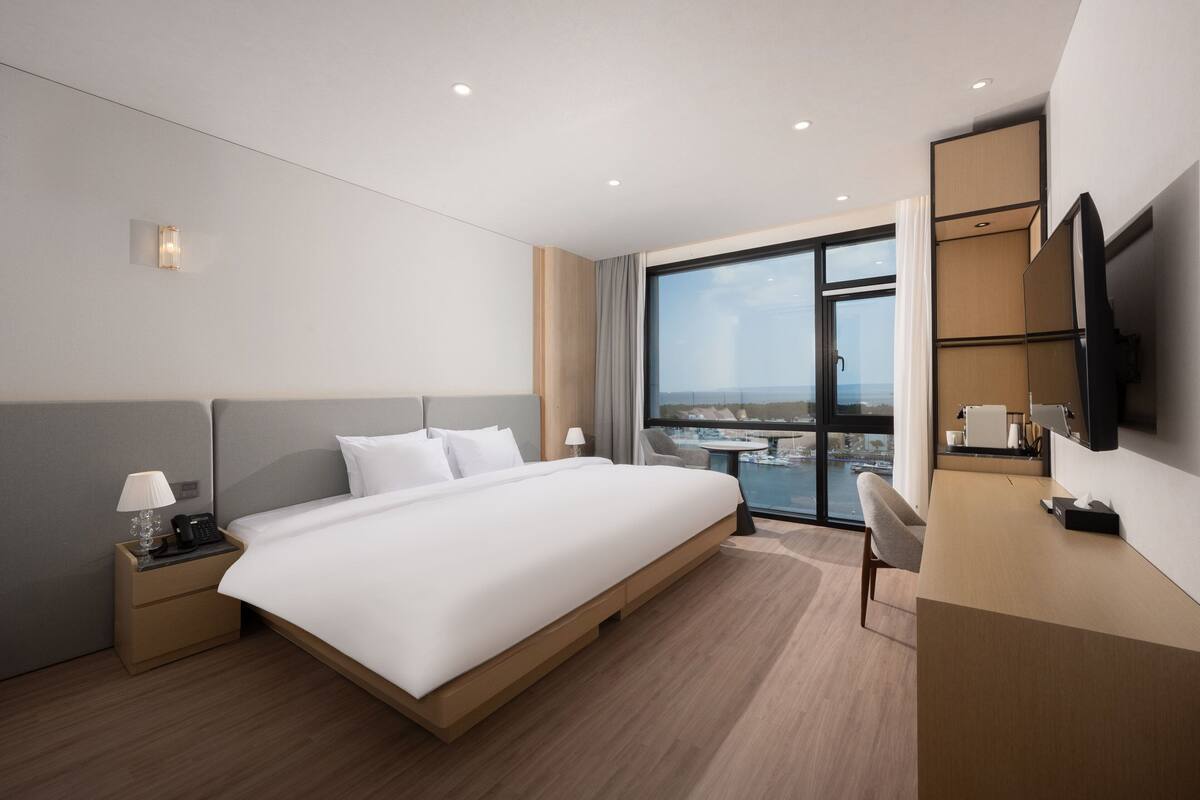 Una habitación de hotel moderna con una cama grande, un escritorio, una silla, un televisor y vistas a la ciudad.