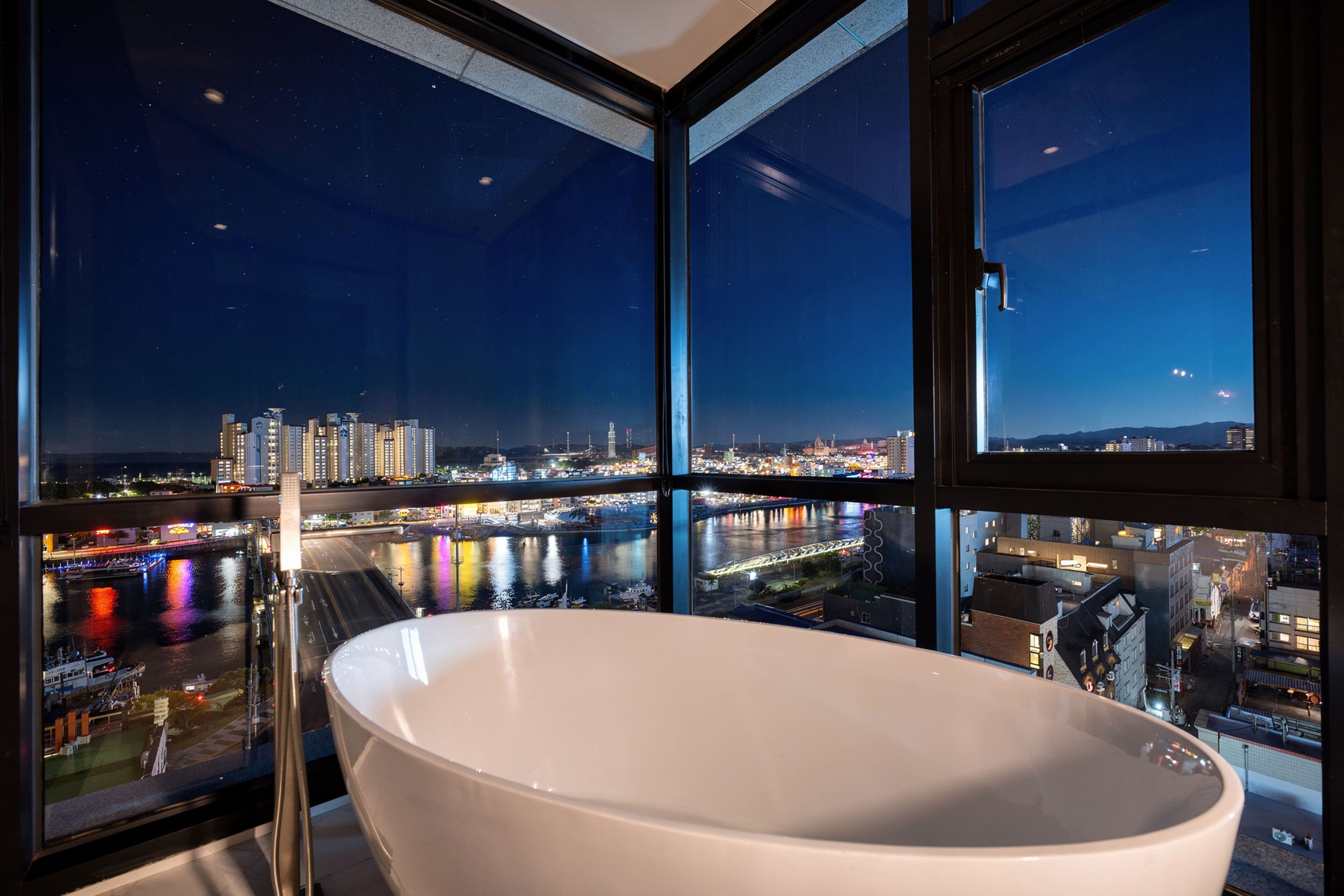 A modern bathroom with a large bathtub, a view of a cityscape at night, and a glass partition.