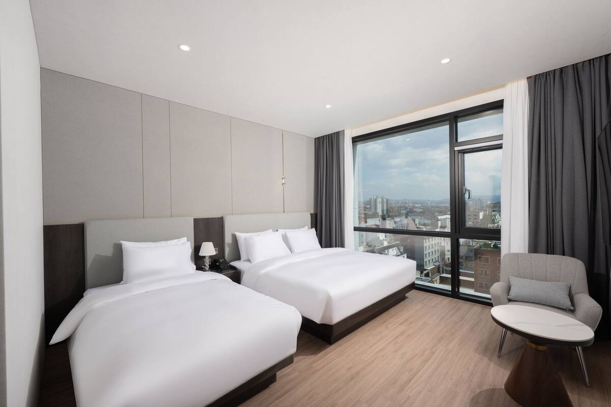 Habitación de hotel con dos camas, una silla, una mesa y un ventanal con vista a la ciudad.