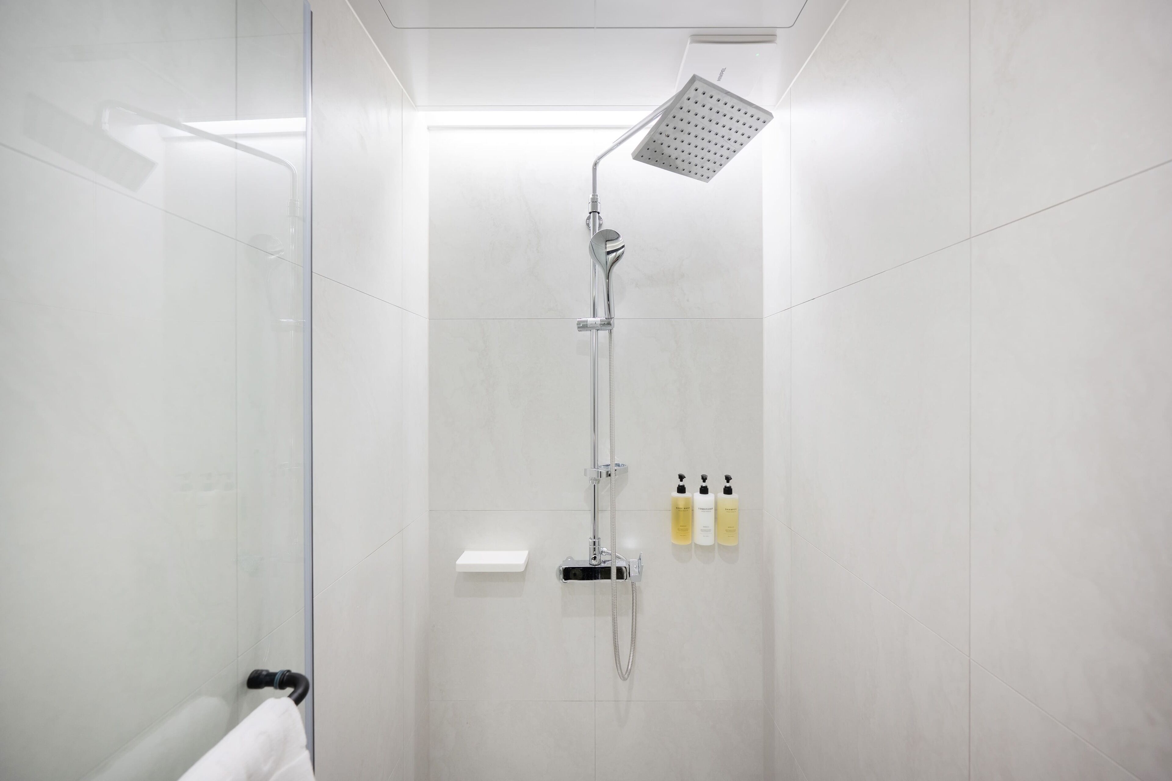 A modern shower with a handheld showerhead and a fixed showerhead, a white towel rack, and three bottles of shampoo and conditioner.