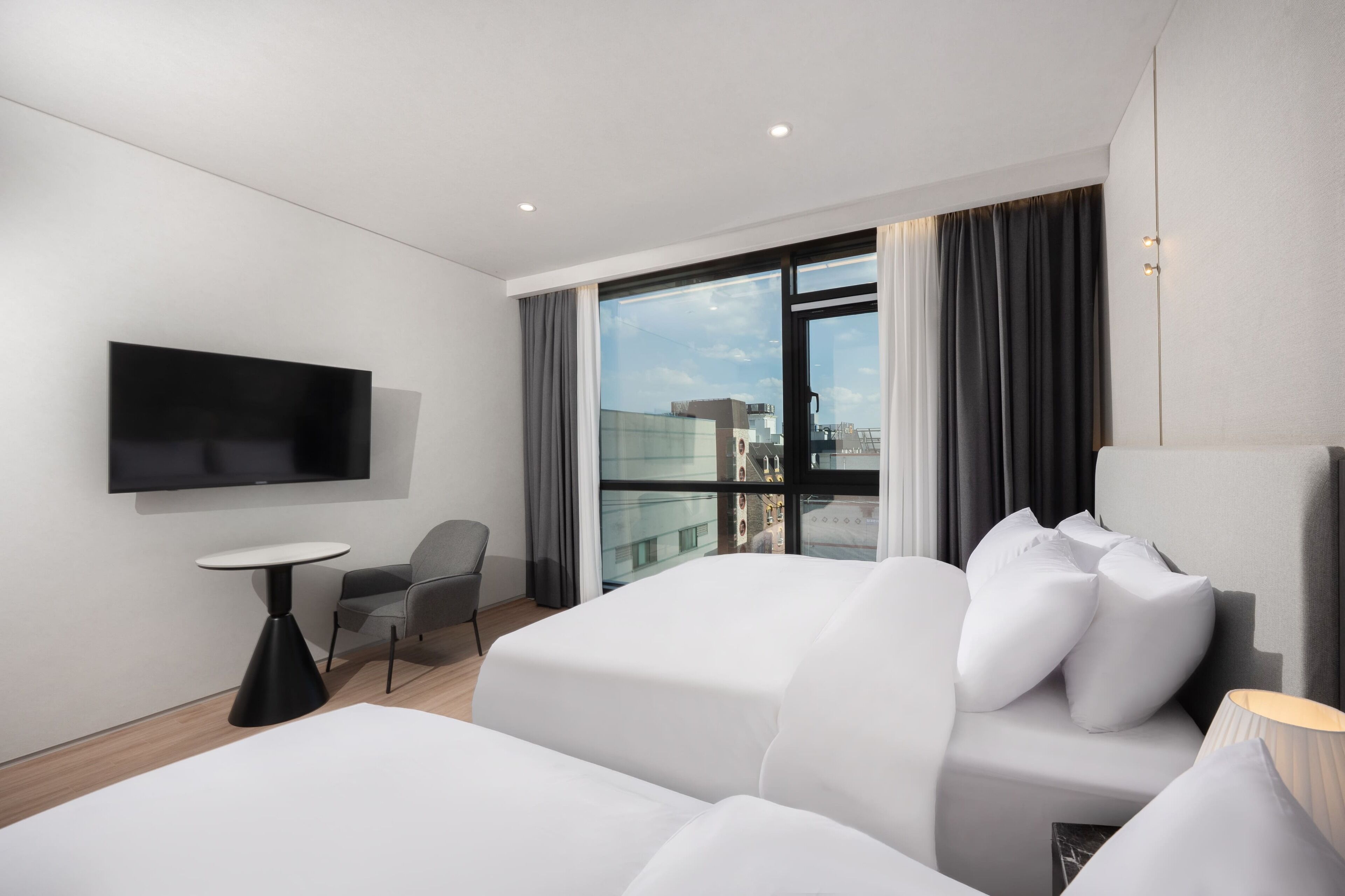 A modern hotel room with a large bed, a TV mounted on the wall, a small table, a chair, and a view of the city through the window.