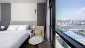 A hotel room with a bed, a chair, a small table, a nightstand, and a view of a cityscape and water.