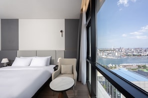 A hotel room with a bed, a chair, a small table, a nightstand, and a view of a cityscape and water. - Pohang City Hotel (Pohang)