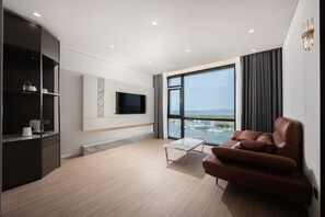Una sala moderna con un sofá, una mesa de centro, un televisor montado en la pared y un amplio ventanal con vistas a un puerto deportivo.