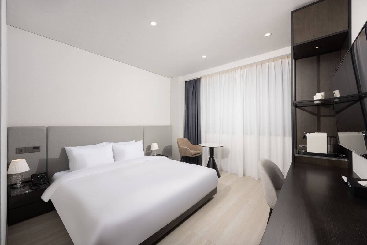 Habitación de hotel con una cama grande, un escritorio, una silla, una mesita, una mesita de noche, una lámpara, un teléfono y un televisor.