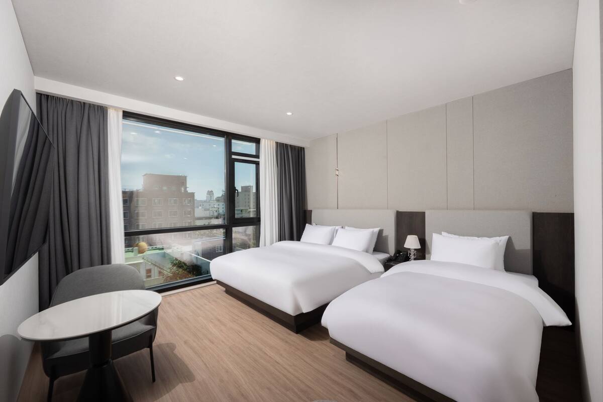 Habitación de hotel con dos camas, una mesita, una silla, una mesita de noche y un ventanal grande con cortinas.