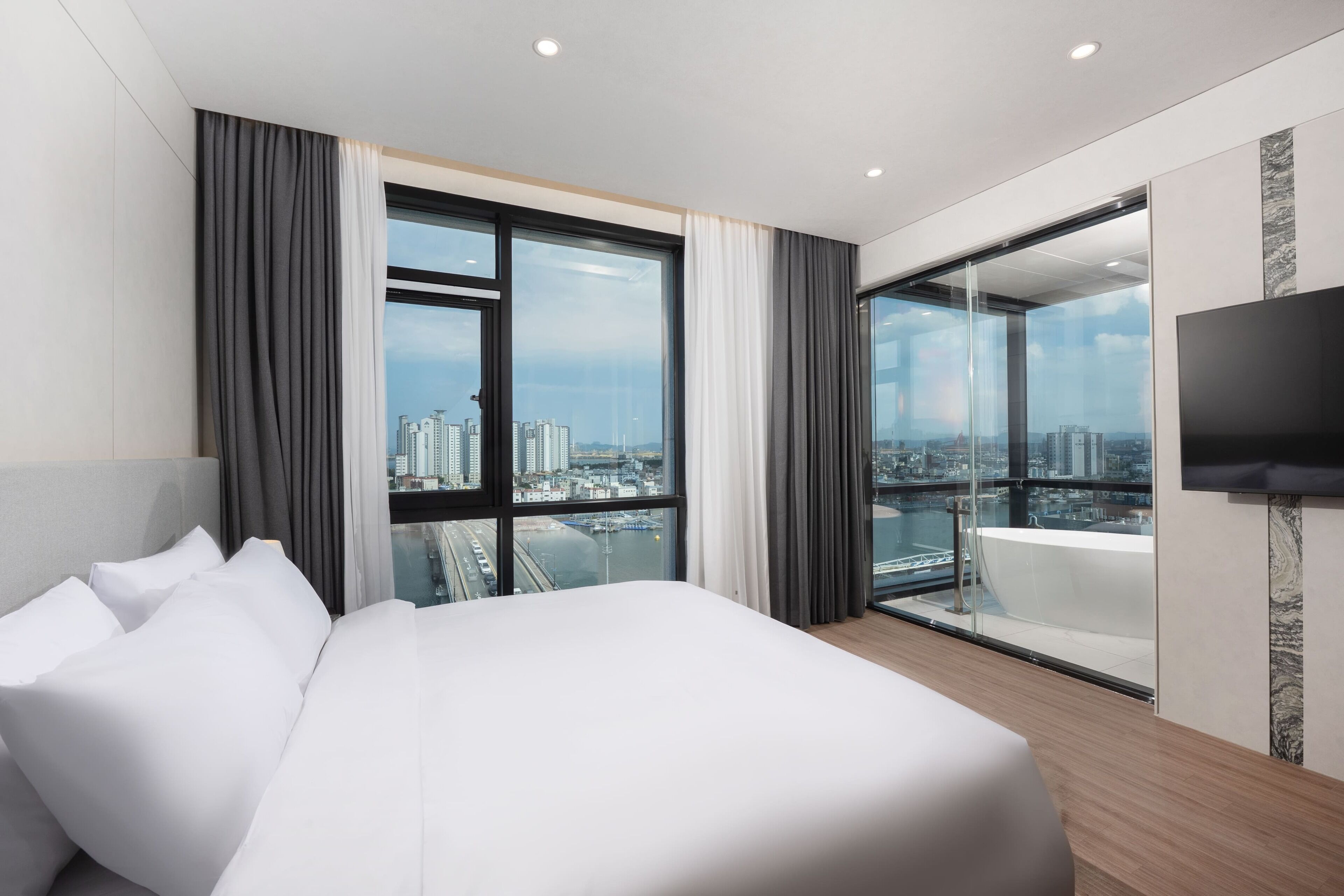 A modern hotel room with a large bed, a city view, a glass door leading to a balcony, and a flat-screen TV mounted on the wall.