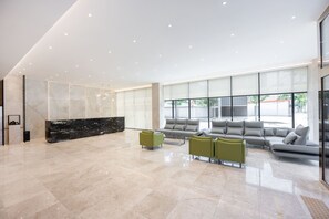 Un hall d’entrée spacieux, doté d’un comptoir de réception en marbre, d’un canapé d’angle gris et d’un mur de verre donnant sur l’extérieur.