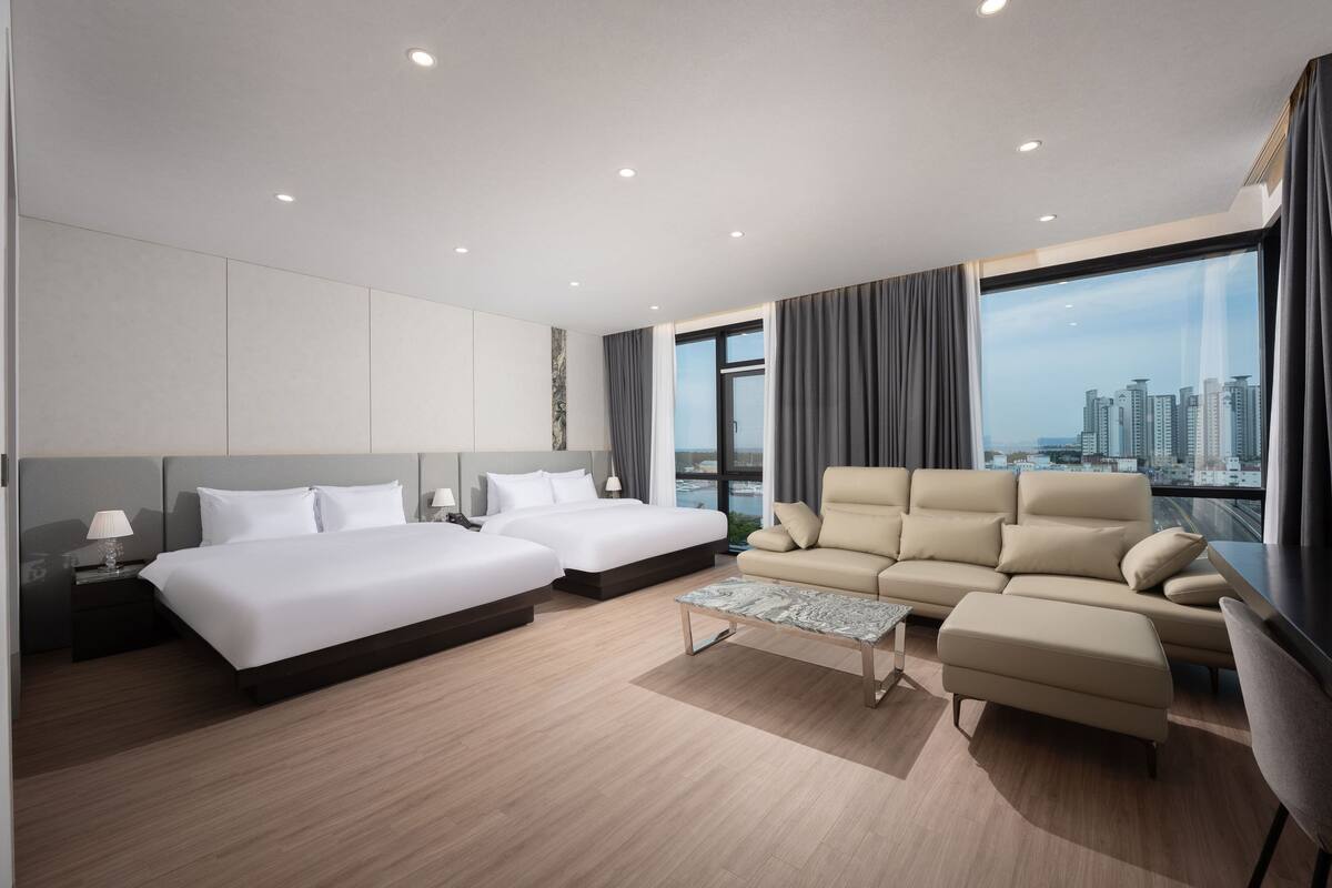 Una habitación de hotel moderna con dos camas, un sofá, una mesa de centro y un ventanal con vistas a la ciudad.