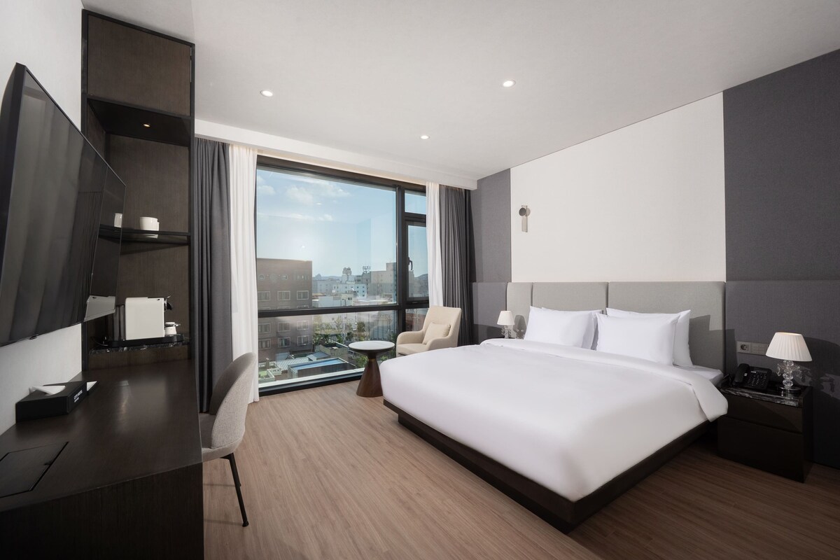Habitación de hotel moderna con una cama grande, un escritorio, una silla, un televisor y vistas a la ciudad.