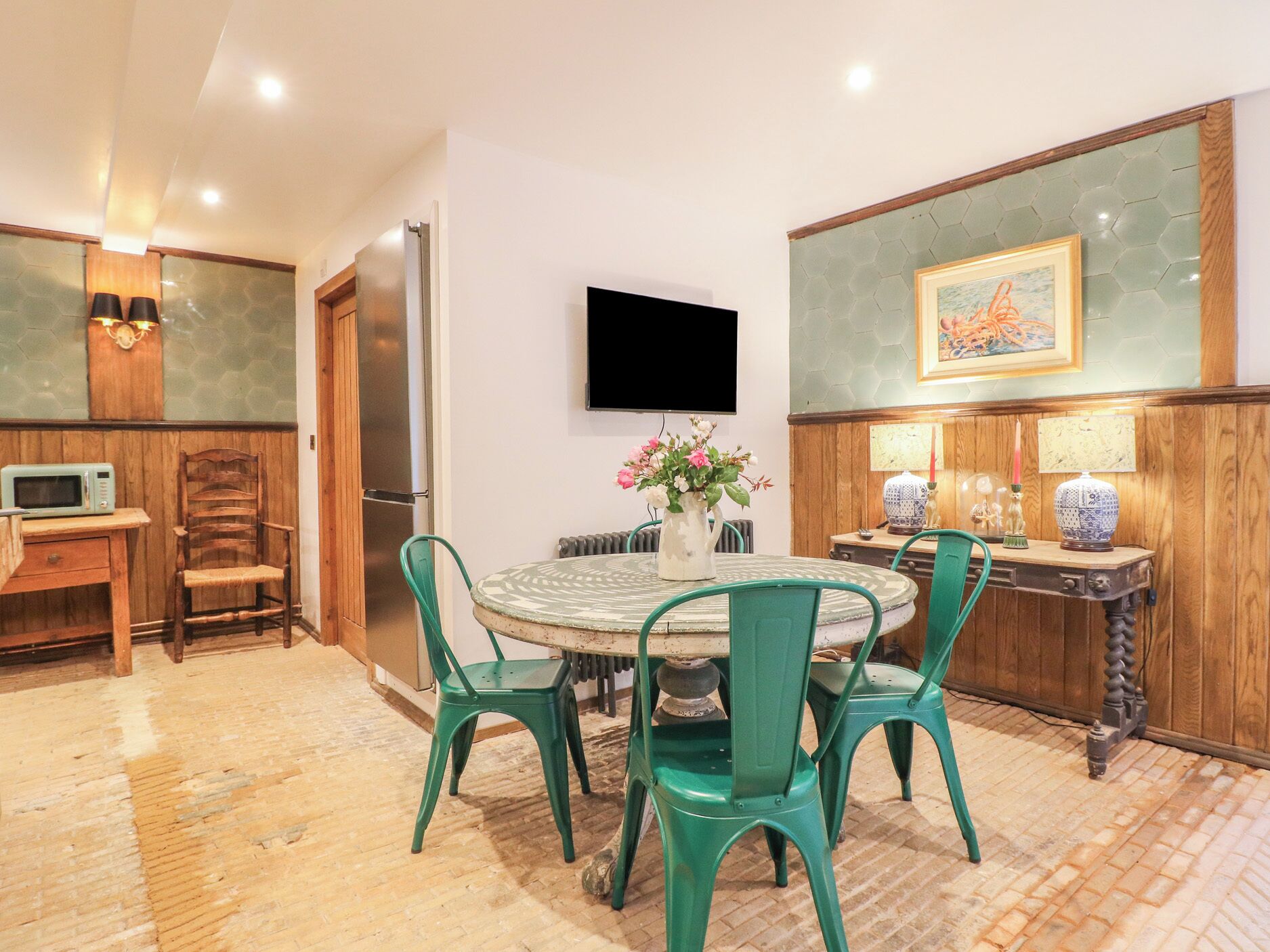 Un coin repas avec une table ronde, des chaises vertes, un vase de fleurs, un téléviseur fixé au mur et un four à micro-ondes posé sur un meuble en bois.