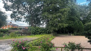A gravel path leading to a fenced area with a wooden gate, surrounded by lush greenery and pink flowers. - Shrewsbury Lodge (Stafford)