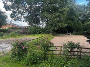 Ein Kiesweg führt zu einem eingezäunten Bereich mit einem Holztor, umgeben von üppigem Grün und rosa Blüten.