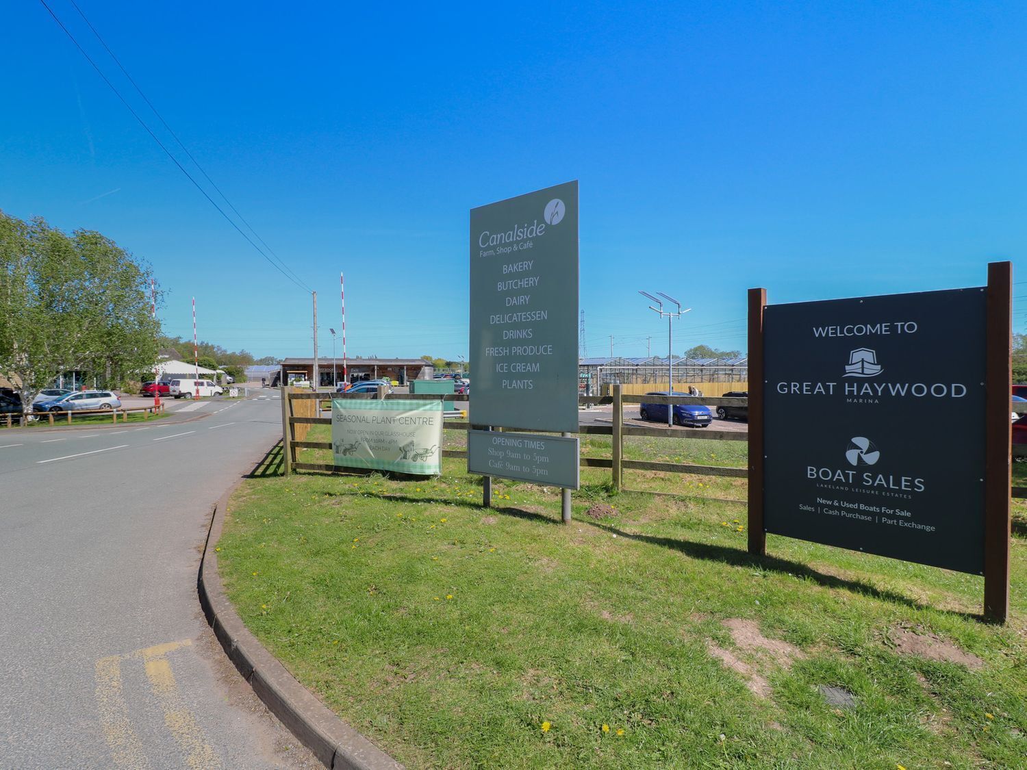 Un cartel al borde de la carretera anuncia un vivero de temporada, con horarios de apertura y un letrero de bienvenida a Great Haywood Marina.