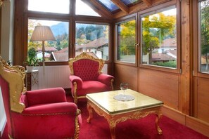 Ein Zimmer mit einem rot gepolsterten Sessel, einem passenden Sofa, einem Glastisch mit einem Weinglas und Blick auf Häuser und Bäume durch große Fenster.
