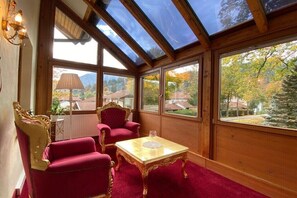 Een zonnedek met glazen dak, twee rode fauteuils, een tafeltje met een glas en uitzicht op een woonwijk.