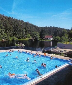 Ein Schwimmbecken mit Personen, umgeben von bewaldeter Landschaft und einem Gebäude im Hintergrund.