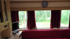 A compact RV interior with a kitchenette, a red sofa, a small table, and a clock on the wall. - Mobile home on a quiet wooded lot with a barbecue and outdoor table. (Couret)