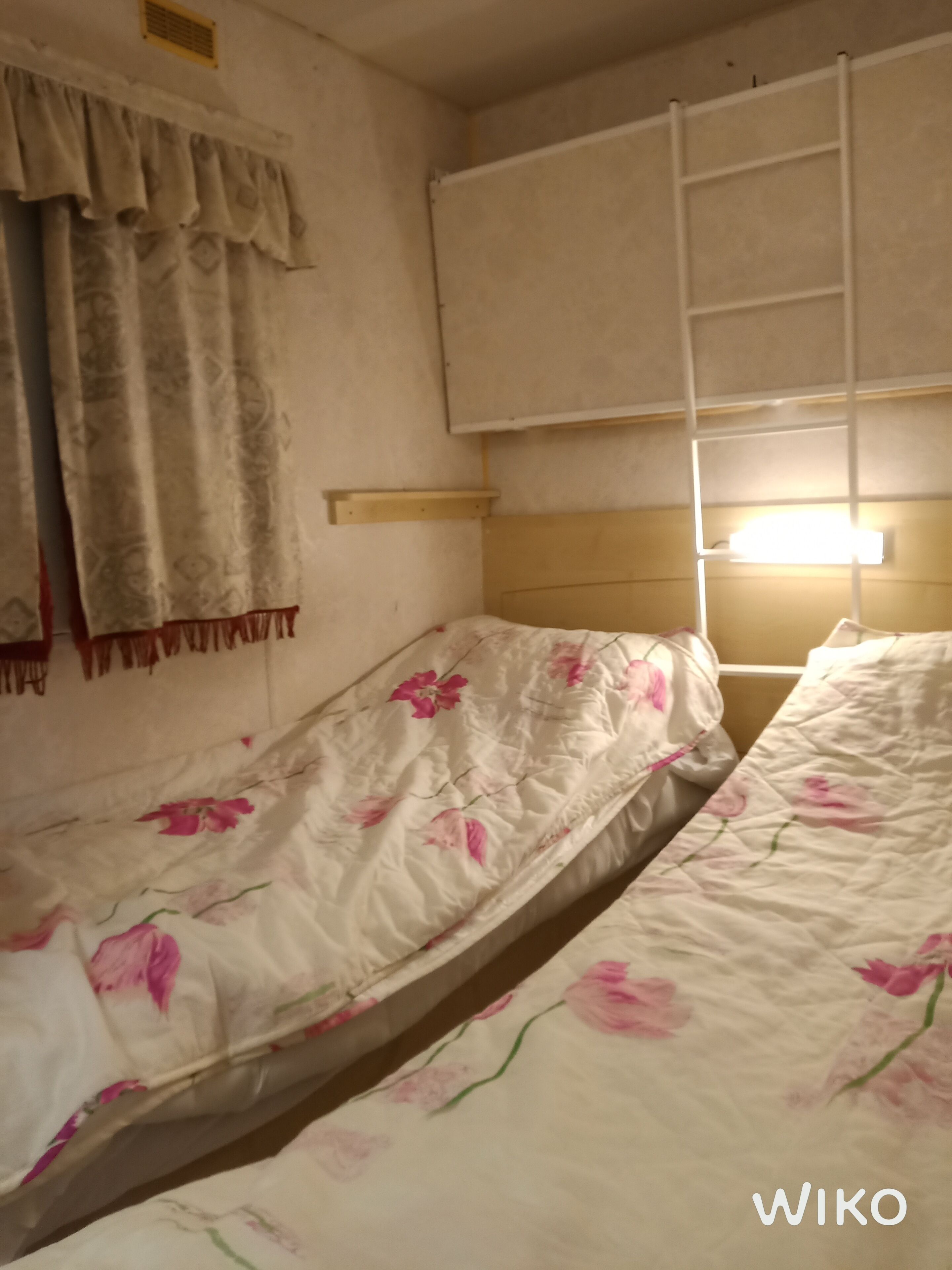 A room with two single beds, a wooden headboard, a window with a white blind, and a wall-mounted shelf.