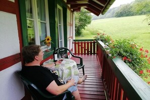 A person seated on a porch with a table and chairs, a window with a sunflower decoration, and a view of a green field. - Apartment in Owingen with swimming pool (Owingen)