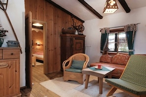 Ein gemütliches Zimmer mit einem Holzkasten, Korbstühlen, einer Couch, einem Tisch und einem Fenster mit Vorhängen.