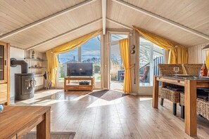 Ein gemütliches Zimmer mit Fernseher, Holzmöbeln, Kamin und großen Fenstern mit Vorhängen.