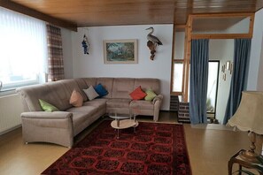 Ein Wohnzimmer mit einem beigefarbenen Ecksofa, einem roten Teppich mit Muster, einem kleinen runden Couchtisch und einem gerahmten Landschaftsbild an der Wand.
