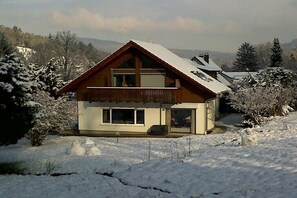 Ein zweistöckiges Holzhaus mit Balkon, umgeben von schneebedeckten Bäumen und Bergen.