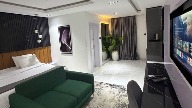 Una habitación de hotel moderna con cama, sofá verde, televisor, escritorio y plantas en macetas.