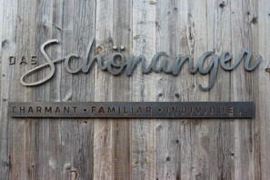Eine rustikale Holzwand mit Metallschildern mit den Aufschriften 'Das Schönanger' und 'CHARMANT • FAMILIÄR • INDIVIDUELL'.