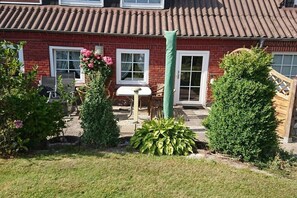 A red brick house with a tiled roof, a garden with various plants, a white table, and a green chair. - Holiday apartment with 2 bedrooms (Steinberg)