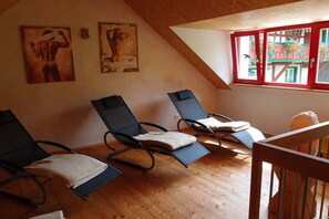 Ein Zimmer mit drei Liegesesseln, Holzboden und einem Fenster mit roten Lamellen.