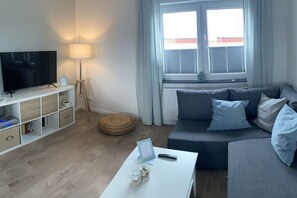 A modern living room with a flat-screen TV, a white coffee table with a remote and a candle, a grey sofa with cushions, a white shelving unit, and a window with blinds. - Holiday apartment Brillau (Kayhude)
