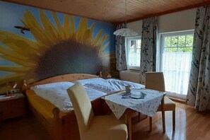 Ein Zimmer mit einem Bett, einem Tisch mit Stühlen, einem Fenster mit Vorhängen und einer Wand mit einem Sonnenblumen-Wandbild.